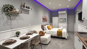 A modern, compact room with a dining area, a bed with a white and yellow bedspread, a small kitchen with a sink, and a mounted TV. - La Casa dell' Arco Nr 45/47 Entire house in historic center (Triggiano)