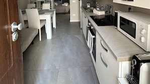 A modern kitchen with white cabinetry, a built-in microwave, and a coffee machine. There is a dining area with a white table and chairs, and a bed in the background. - La Casa dell' Arco Nr 45/47 Entire house in historic center (Triggiano)