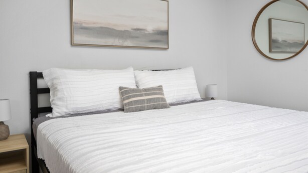 A neatly made bed with white bedding and a gray striped pillow, a bedside lamp, and a framed picture on the wall. - Inn at Rockaway Beach #1: Walk to Beach, Pets OK (Rockaway Beach)