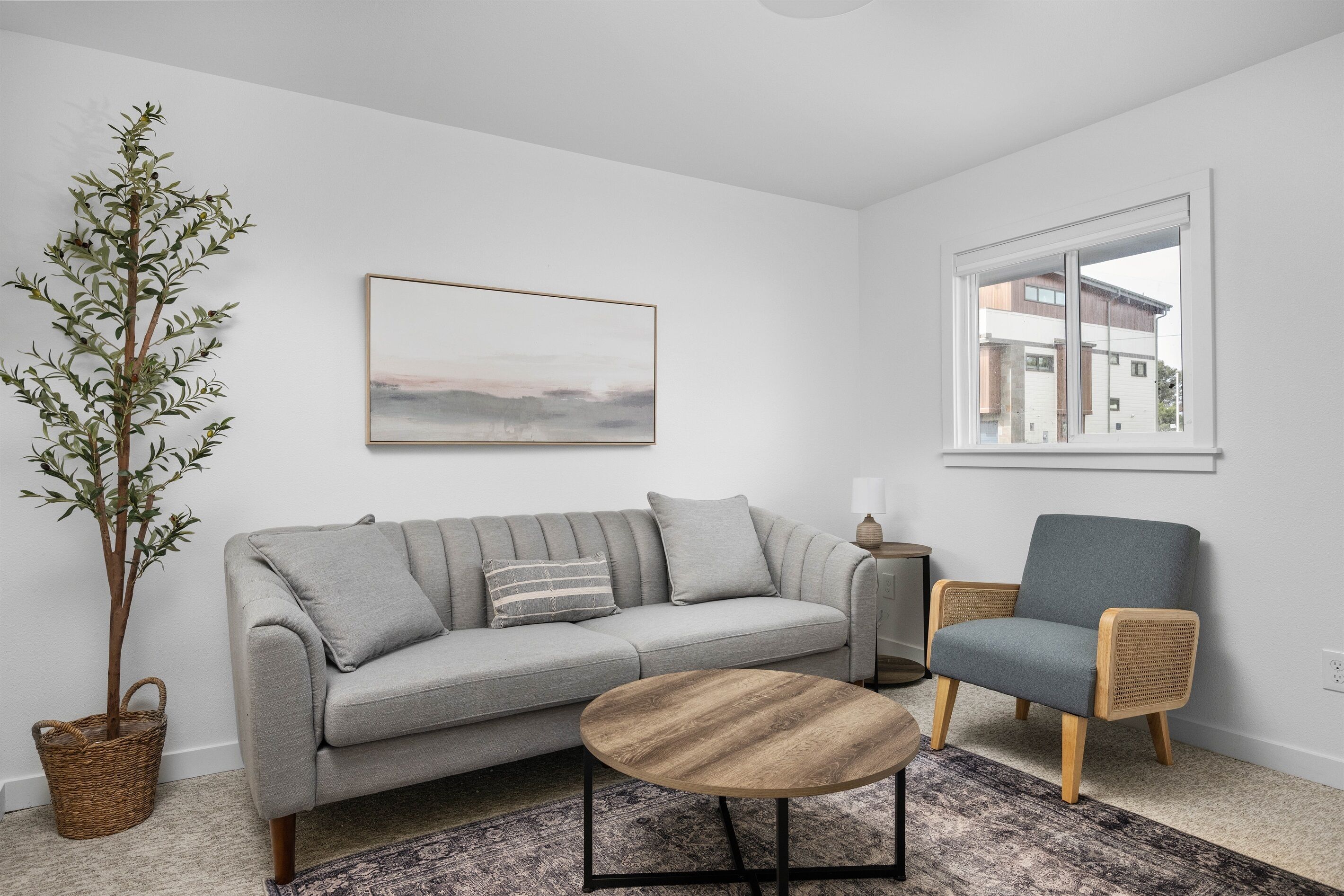 A modern living room with a grey sofa, a round wooden coffee table, a wicker chair, a potted plant, and a framed picture on the wall.