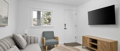 A modern living room with a grey sofa, a wooden coffee table, a grey armchair, a wooden TV stand, and a large flat-screen TV.