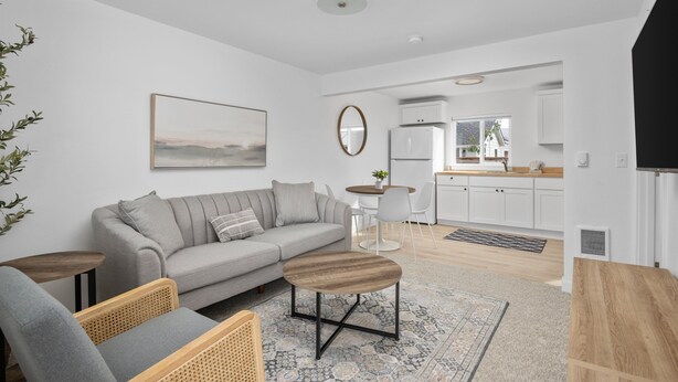 Ett modernt vardagsrum med en grå soffa, ett soffbord och en matplats med ett litet bord och stolar. I bakgrunden syns ett kök med vita skåp och ett kylskåp.