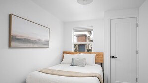 Ein ordentlich bezogenes Bett mit hölzernem Kopfteil, ein Fenster mit Blick auf Gebäude und eine schlichte weiße Tür.