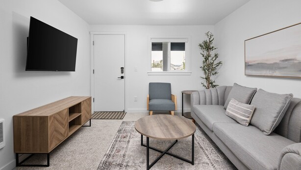 A modern living room with a flat-screen TV, a wooden TV stand, a grey sofa with cushions, a round coffee table, and a window with a view. - Inn at Rockaway Beach #9: Walk to Beach, Pets OK (Rockaway Beach)