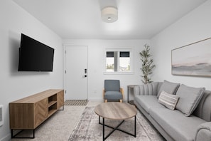 A modern living room with a flat-screen TV, a wooden TV stand, a grey sofa with cushions, a round coffee table, and a window with a view. - Inn at Rockaway Beach #9: Walk to Beach, Pets OK (Rockaway Beach)