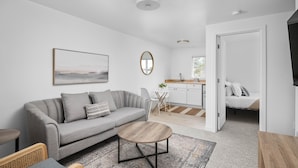 A modern living room with a grey sofa, a round coffee table, and a dining area with a kitchen in the background. - Inn at Rockaway Beach #9: Walk to Beach, Pets OK (Rockaway Beach)