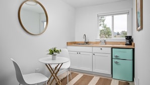 A compact kitchen with white cabinets, a small dining table with chairs, a round mirror, a window with a view, and a retro-style refrigerator. - Inn at Rockaway Beach #9: Walk to Beach, Pets OK (Rockaway Beach)