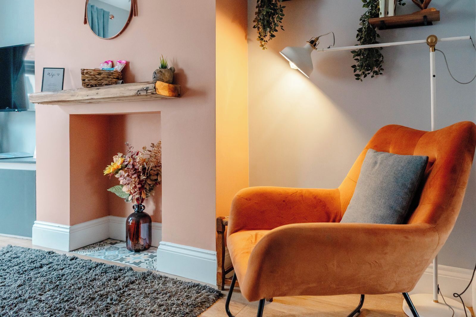A room with a wooden shelf, a vase with flowers, a chair with a cushion, and a floor lamp.