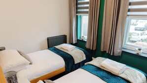 Een hotelkamer met twee bedden, een raam met gordijnen, een radiator en een kleine plant op de vensterbank.