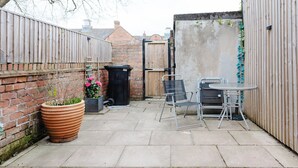 Een terras met een bakstenen muur, een metalen tafel en stoelen, een grote terracotta pot met planten en een zwarte afvalbak.