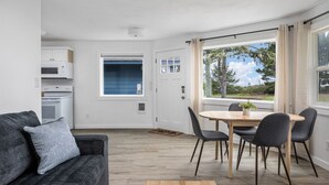 Uma sala de estar moderna com sofá, mesa de jantar com cadeiras, mesa de centro, área de cozinha com eletrodomésticos e uma grande janela com vista para árvores e céu.