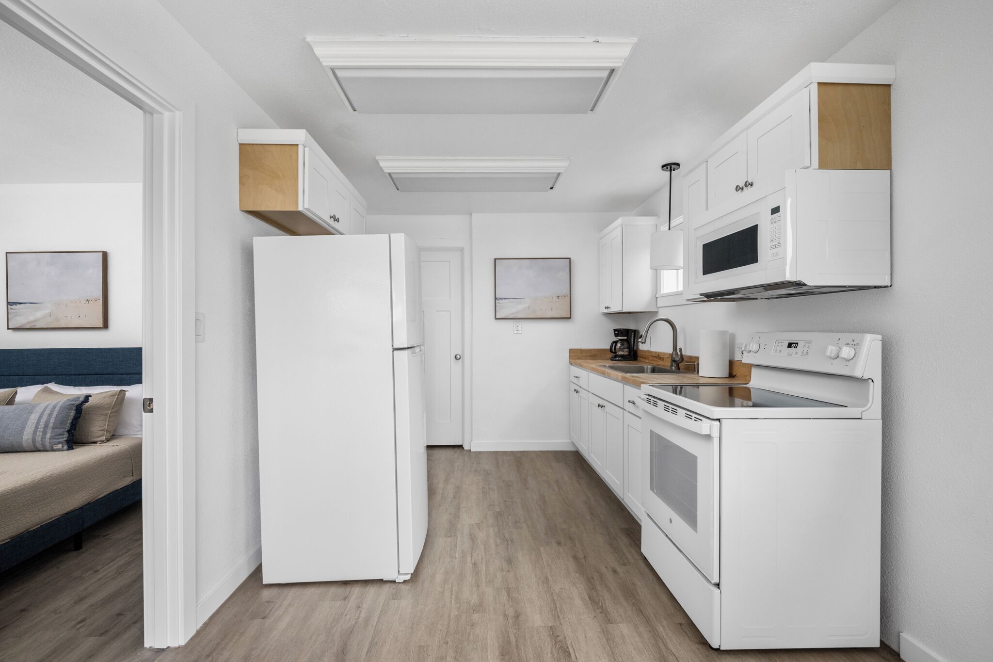 Eine moderne Küche mit weißen Geräten, einer Holz Arbeitsplatte und Blick in ein Schlafzimmer mit Bett und einem Wandbild.
