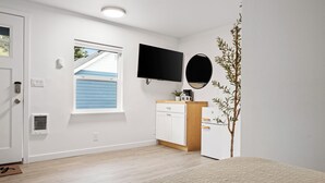 A room with a white door, a window with blue shutters, a flat-screen TV mounted on the wall, a wooden cabinet with a mirror, and a small plant. - Ocean Rogue Inn Suite #10 - Suite | Oceanfront (Rockaway Beach)