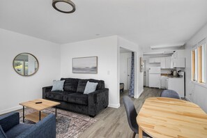 A modern living room with a dark grey sofa, a wooden coffee table, and a blue armchair. A kitchen with white cabinets and a dining area with a wooden table and chairs is visible in the background. - Ocean Rogue Inn Suite #2 - 1 BD Condo | Oceanfront (Rockaway Beach)