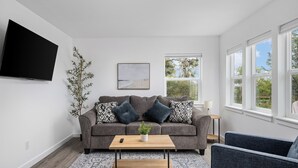 Ein modernes Wohnzimmer mit grauem Sofa, einem Couchtisch, einem an der Wand montierten Fernseher und einem Fenster mit Blick auf Grünflächen.