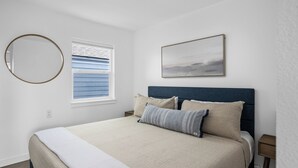 A bedroom with a bed, a round mirror, a window with blinds, and a framed picture on the wall. - Ocean Rogue Inn Suite #4 - 1 BD Condo | Oceanfront (Rockaway Beach)