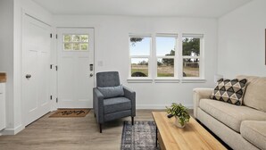 Un salon avec un fauteuil gris, un canapé beige, une table basse en bois ornée d’une plante, et une vue sur l’extérieur grâce à de grandes fenêtres.