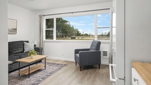 Een moderne woonkamer met een grijze bank, een houten salontafel, een grijze fauteuil en een groot raam met uitzicht op bomen en een heldere lucht.