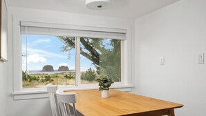 A room with a wooden table and white chairs, a window with a view of the beach, and a potted plant on the table.