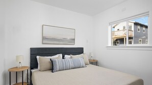 A bedroom with a large bed, two bedside tables, a nightstand, a window with a view of a building, and a framed picture on the wall.