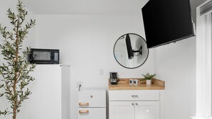 Cozinha compacta com geladeira branca, micro-ondas, cafeteira e um pequeno armário. Há uma planta em vaso sobre o balcão e uma TV de tela plana fixada na parede.