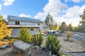 A two-story house with a metal roof, surrounded by a landscaped garden with various plants and a stone bench. - The Swell House: Coastal Luxury & Private En-Suites (Pacific City)