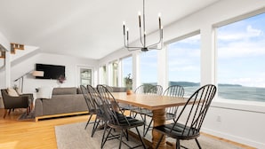 Un vaste espace de vie comprenant une table à manger, des chaises et un canapé. Une grande fenêtre offre une vue sur l’océan.