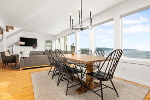 A spacious living area with a dining table, chairs, and a sofa. There is a large window offering a view of the ocean. - Beautiful Oceanside Retreat: 7 Bedrooms, Hot Tub, Views, Sleeps Big Groups (Oceanside)