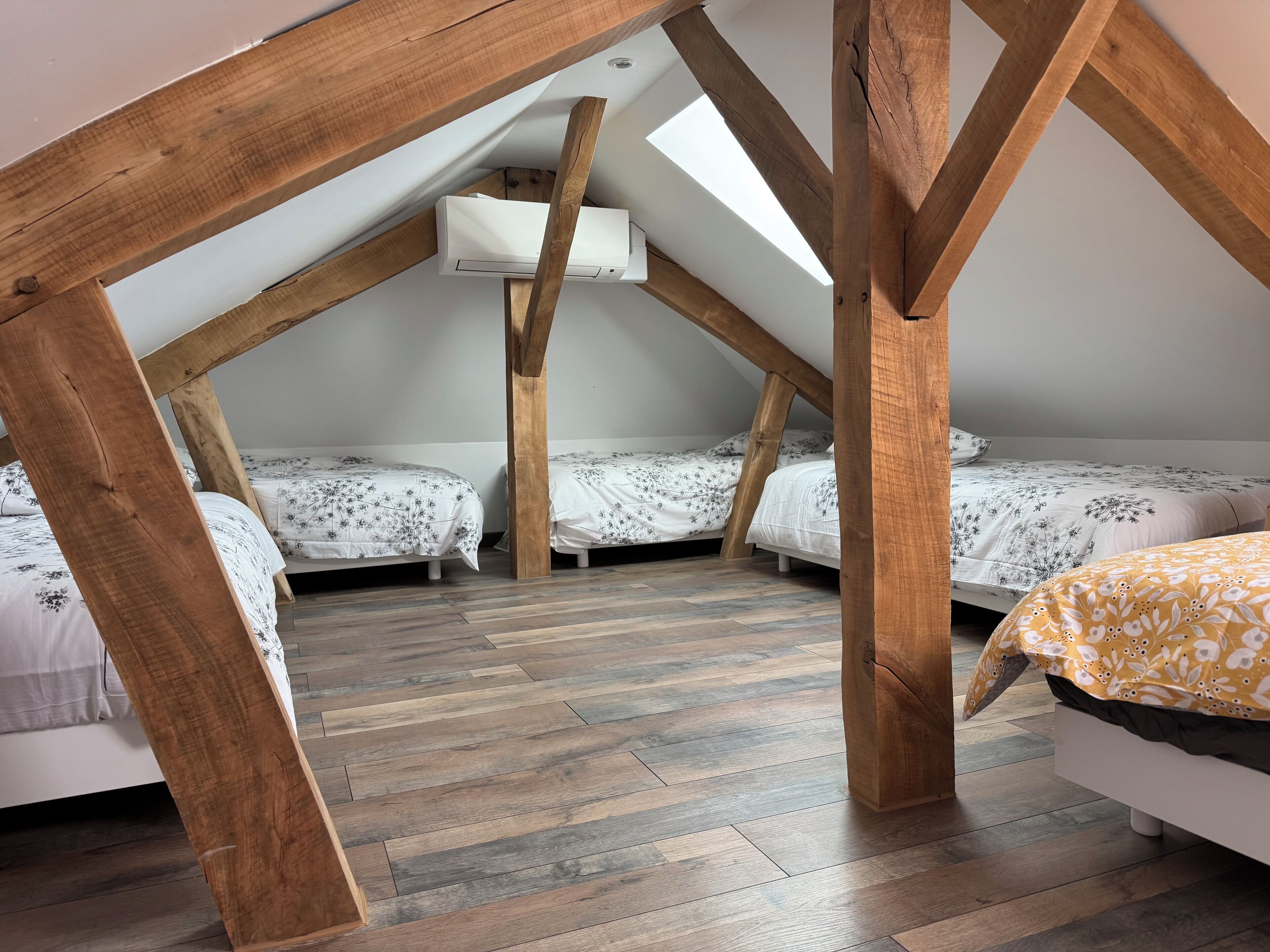 A loft bedroom with exposed wooden beams, three beds with floral bedding, and a skylight.
