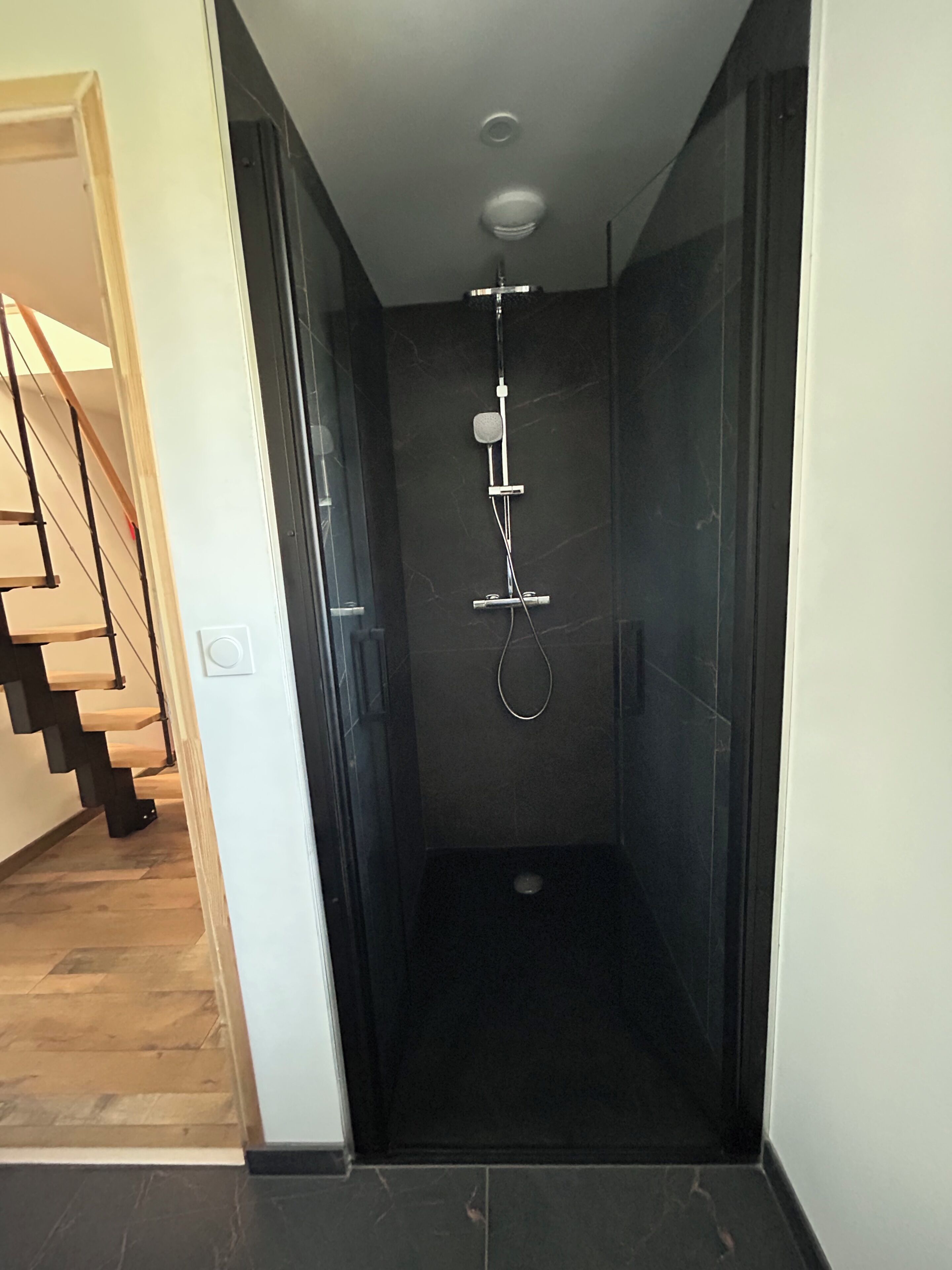 Ein modernes Badezimmer mit einer gläsernen Dusche, einem sichtbaren Lichtschalter und einer Holztreppe im Hintergrund.