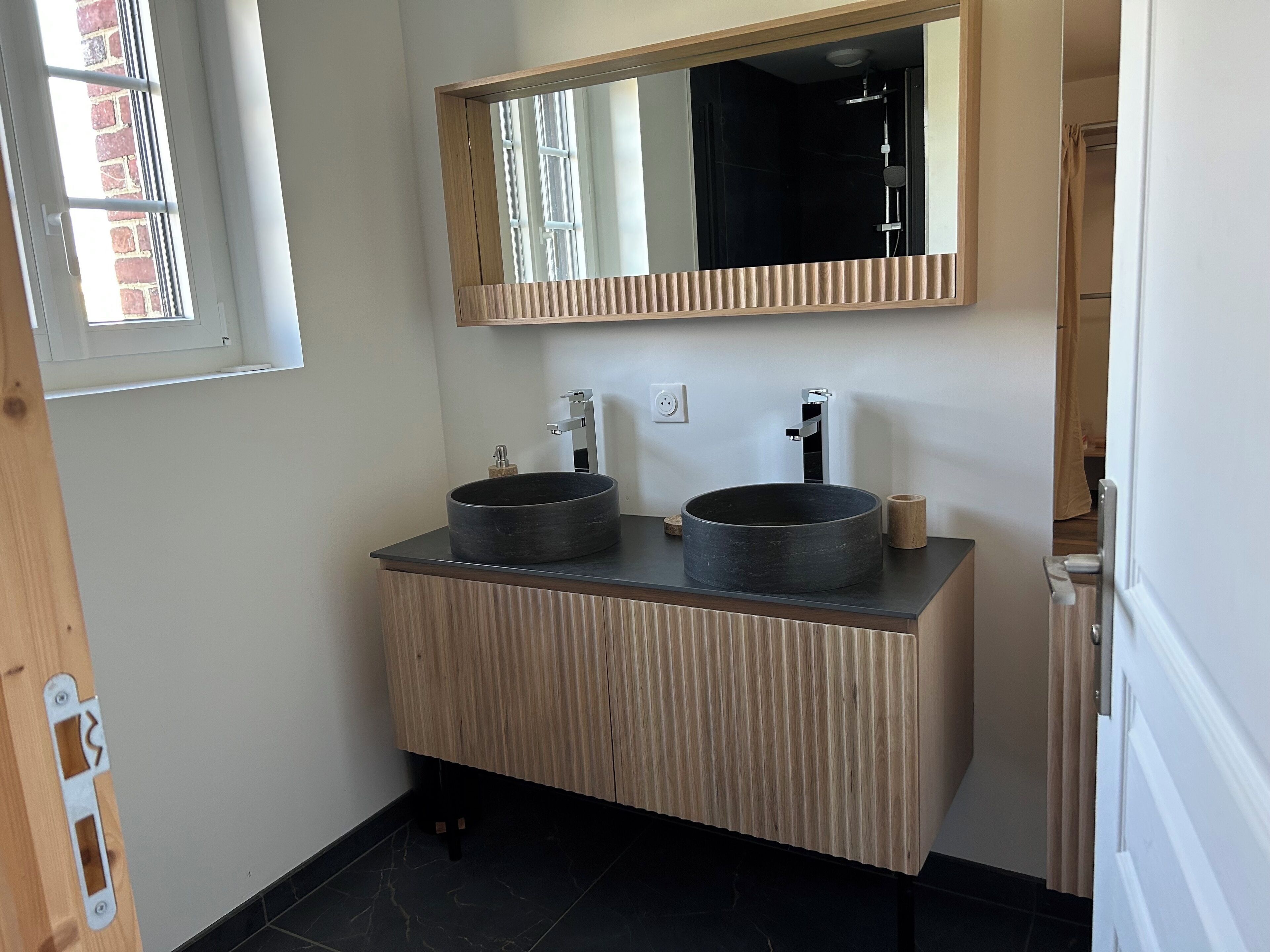 A bathroom with a wooden vanity, two round black sinks, a mirror, and a window with a view of a brick building.