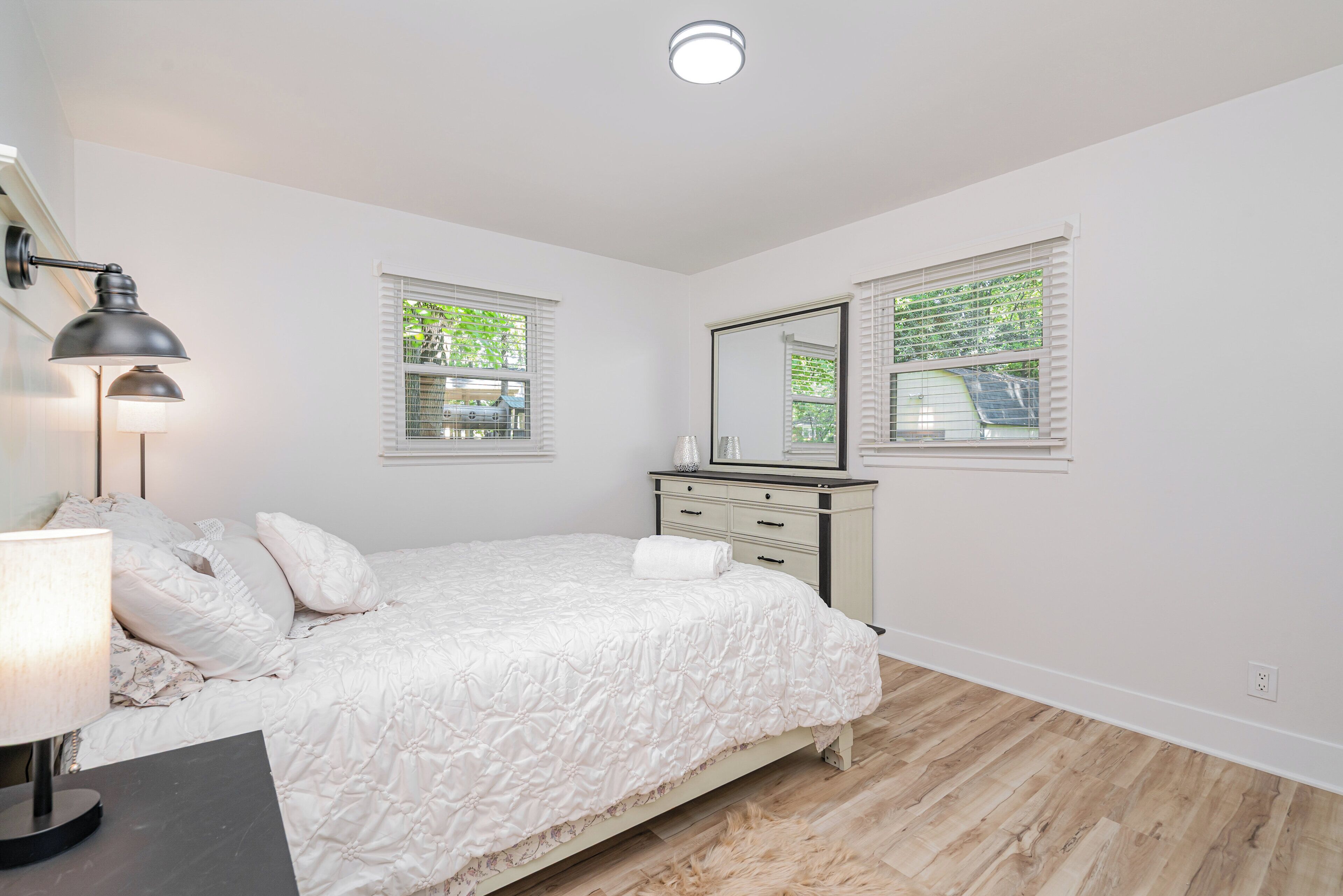 A bedroom with a bed, bedside table, lamp, dresser, and two windows with blinds.