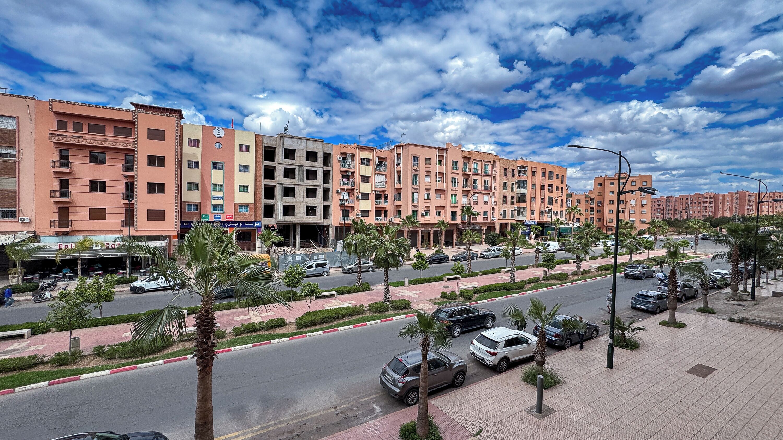 A multi-story residential building with balconies, a street with cars, and palm trees.