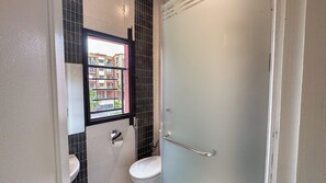 Une salle de bain comprenant des toilettes, un lavabo, une fenêtre donnant sur des immeubles et une porte de douche.