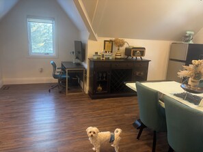 Una habitación con piso de madera, un escritorio con computadora, una silla, una mesa de comedor con sillas, un armario y un perro pequeño.
