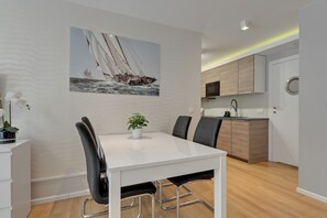 A modern dining area with a white table, black chairs, and a potted plant. A sailboat painting adorns the wall. - Szafarnia 1 by Grand Apartments (Gdańsk)