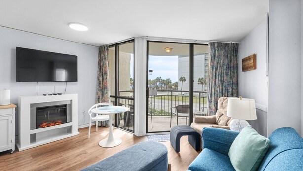 A modern living room with a flat-screen TV, a fireplace, a dining area, and a balcony with a view of palm trees. - One bedroom ocean view Meridian plaza (Myrtle Beach)