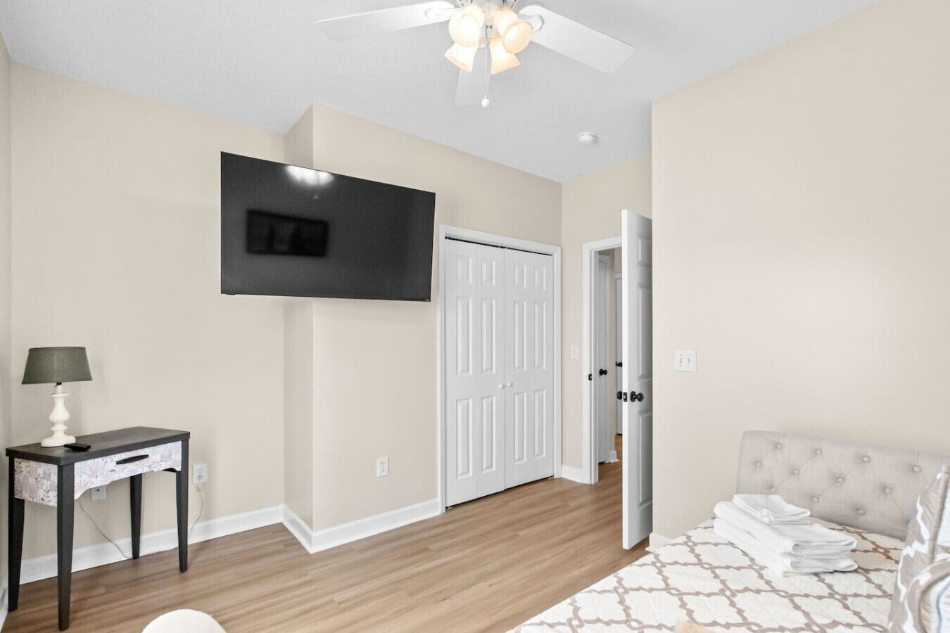 A bedroom with a bed, a bedside table with a lamp, a ceiling fan, a TV mounted on the wall, and a door leading to another room.