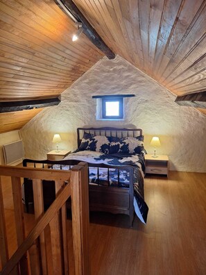 Un dormitorio acogedor en el ático, con techo inclinado, un tragaluz pequeño, una cama de madera con colcha estampada, dos lámparas de noche y una silla de madera.