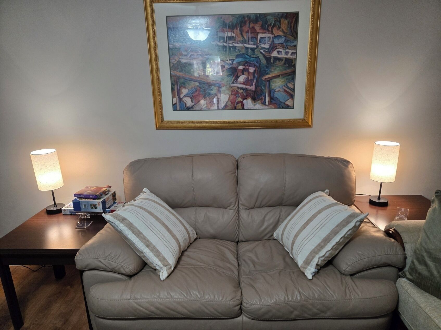 Een woonkamer met een beige bank, twee lampen, een lijst met een schilderij, en een tafeltje met boeken en een lamp.