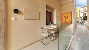 A balcony with a glass railing, a small table with chairs, a wall-mounted light switch, and a painting of a cat. - Your urban oasis in the heart of Chania Town,a blend of convenience and comfort. (Chania)
