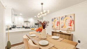 A modern kitchen with white cabinets, a wooden dining table set with plates and a vase of flowers, and abstract art on the wall. - Spacious 4BR Home - King Suite - Family Friendly - Near Dallas Attractions (Dallas)