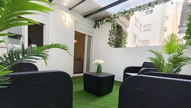 Un área de estar exterior moderna con mobiliario de ratán negro, césped artificial verde y plantas en macetas.