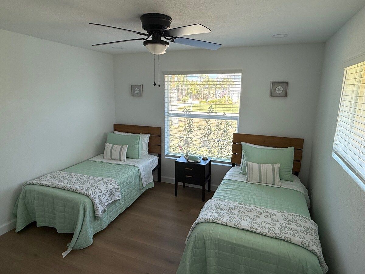 A room with two single beds, a ceiling fan, a small table, and a window with blinds.