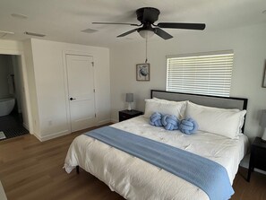 A bedroom with a bed, a ceiling fan, a window with blinds, a bedside table with a lamp, and a door leading to another room. - Beautiful sanibel home (Sanibel)