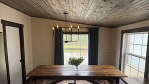A room with a wooden table and bench, a hanging light fixture, a window with curtains, and a door. - NEW! Spacious Farmhouse Cabin, 30 acres w/ Firepit, Pond, Waterfall, Trails (Zoe)