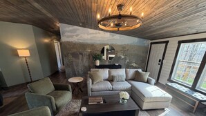 A living room with a sectional sofa, a round coffee table, and a large window. - NEW! Spacious Farmhouse Cabin, 30 acres w/ Firepit, Pond, Waterfall, Trails (Zoe)