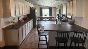 A kitchen with wooden cabinets, a central island with bar stools, a refrigerator, and a window with blue curtains. - NEW! Spacious Farmhouse Cabin, 30 acres w/ Firepit, Pond, Waterfall, Trails (Zoe)
