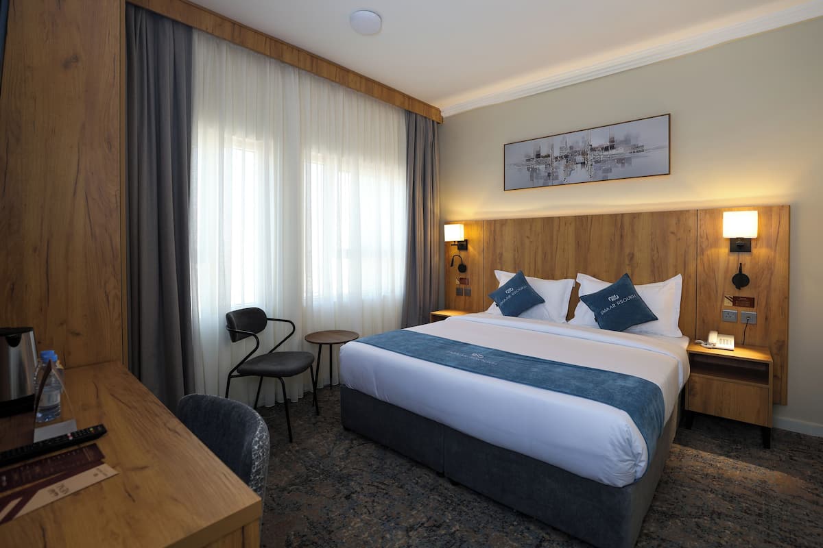 Une chambre d’hôtel avec un grand lit, une tête de lit en bois, un bureau avec une chaise, une petite table et une lampe fixée au mur.