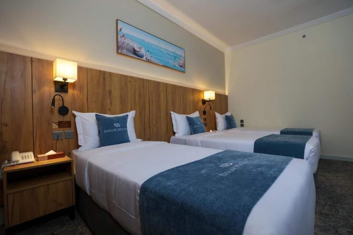 Une chambre d’hôtel avec deux lits, une tête de lit en bois, des lampes fixées au mur, un tableau représentant une scène de plage et un téléphone sur la table de chevet.